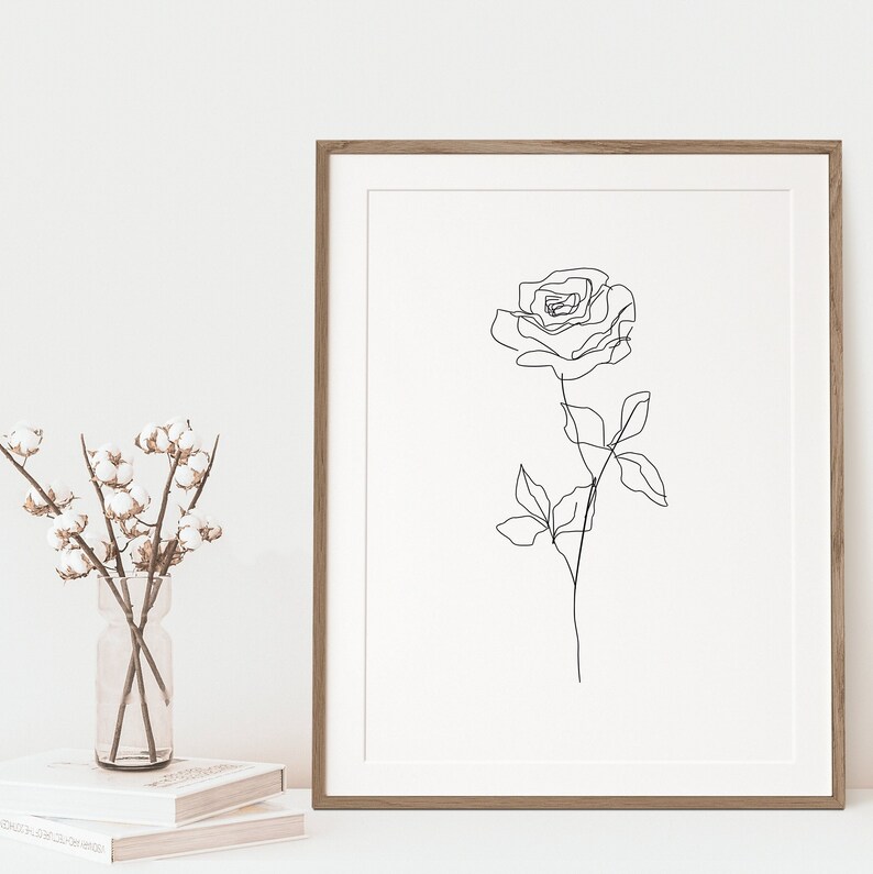 May include: A framed art print featuring a minimalist line drawing of a rose. The artwork is in black against a white background, encased in a wooden frame. A vase of cotton stems and a stack of books are in the foreground.