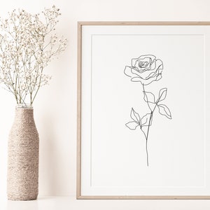 May include: A framed minimalist art print featuring a single-line drawing of a rose. The artwork is in black against a cream background, encased in a light wood frame. A vase wrapped in twine holds white flowers.
