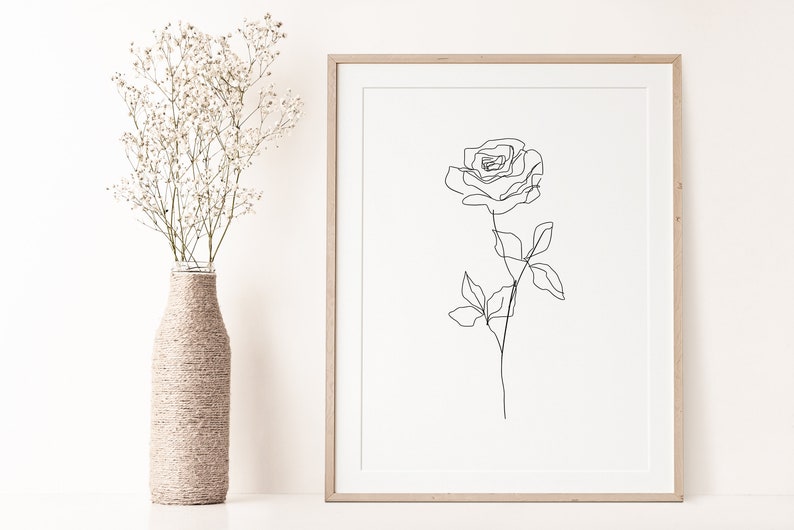 May include: A framed minimalist art print featuring a single-line drawing of a rose. The artwork is in black against a cream background, encased in a light wood frame. A vase wrapped in twine holds white flowers.