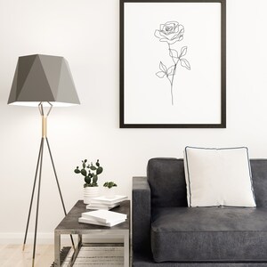 May include: A modern living room scene featuring a black framed art print of a rose outline. A gray geometric lamp, a dark gray sofa with a white pillow, and a metal side table with books and plants complete the decor.