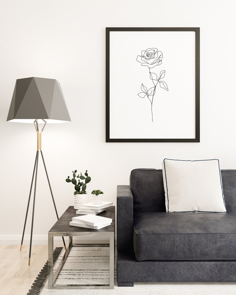 May include: A modern living room scene featuring a black framed art print of a rose outline. A gray geometric lamp, a dark gray sofa with a white pillow, and a metal side table with books and plants complete the decor.
