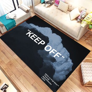 Può includere: Un tappeto rettangolare nero con le parole "KEEP OFF" in grandi lettere bianche. Un design a forma di nuvola in tonalità di grigio e bianco riempie il lato destro. Il tappeto include anche il testo in basso: "IKEA c/o Virgil Abloh Flooring Almhult, Sweden c.2019."