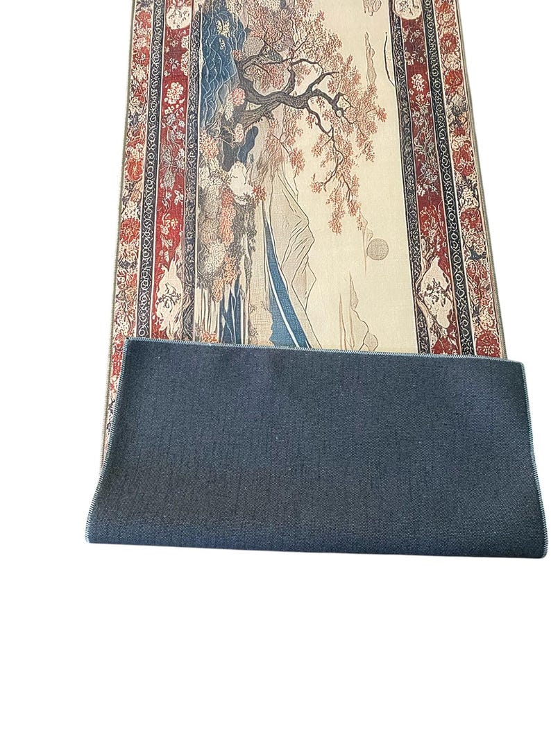 May include: A long, rectangular rug with a cream-colored background and a detailed, colorful design. The rug features a traditional Japanese landscape scene with a tree, mountains, and water. The rug has a dark blue border with red and gold accents.