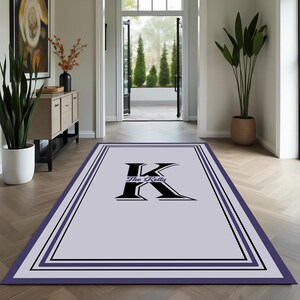 May include: A rectangular, light purple rug with a black and dark purple border. The rug features a large black letter "K" with the words "The Kelly" underneath. The rug is placed on a wooden floor in a hallway.