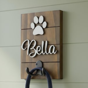 May include: A rectangular wooden wall hook featuring a dark brown finish with horizontal slats. The board displays a white paw print icon above the white script text Bella. A black metal hook is attached, holding a dark blue pet leash.