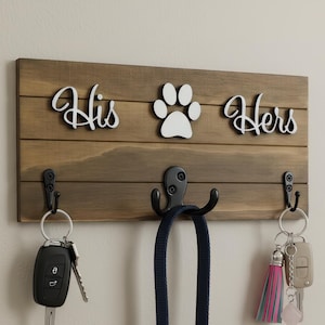 May include: A rectangular wooden wall-mounted key and leash holder with three black metal hooks. The board features white cutout lettering that reads His, a paw print icon, and the word Hers. Keys hang from the outer hooks, and a leash hangs from the center hook.