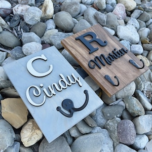 May include: Two wooden signs with hooks for hanging items. The sign on the left is gray with white lettering that says "Cindy" and a black hook. The sign on the right is brown with black lettering that says "Martin" and two black hooks.