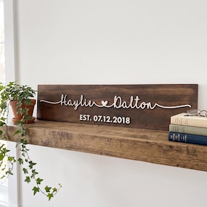 May include: A rectangular dark brown wooden sign featuring white 3D lettering that reads Haylie and Dalton with a small heart symbol in between. Below the names, the text reads EST. 07.12.2018 in white. The sign is displayed on a wooden shelf.