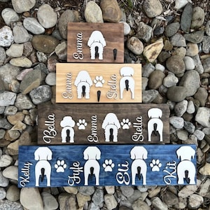 May include: Wooden dog leash holders in various sizes and colors. Each holder features a white dog silhouette, paw prints, and personalized names. The holders are mounted on a brown, tan, and blue background.