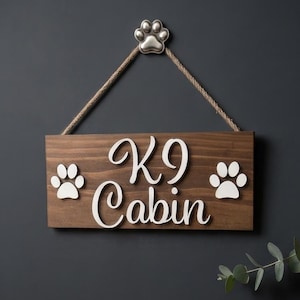 May include: A rectangular wooden sign with a dark brown finish featuring the white text K9 Cabin and two white paw prints. The sign hangs from a jute rope attached to a silver-coloured metal paw print accent at the top centre of the display.