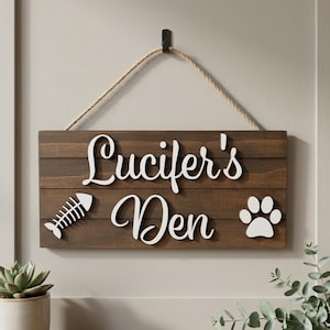 May include: A rectangular dark brown wooden sign hanging from a rope. The sign features white raised lettering that reads "Lucifer's Den," accompanied by a white graphic of a fish skeleton on the left and a white paw print on the right side.