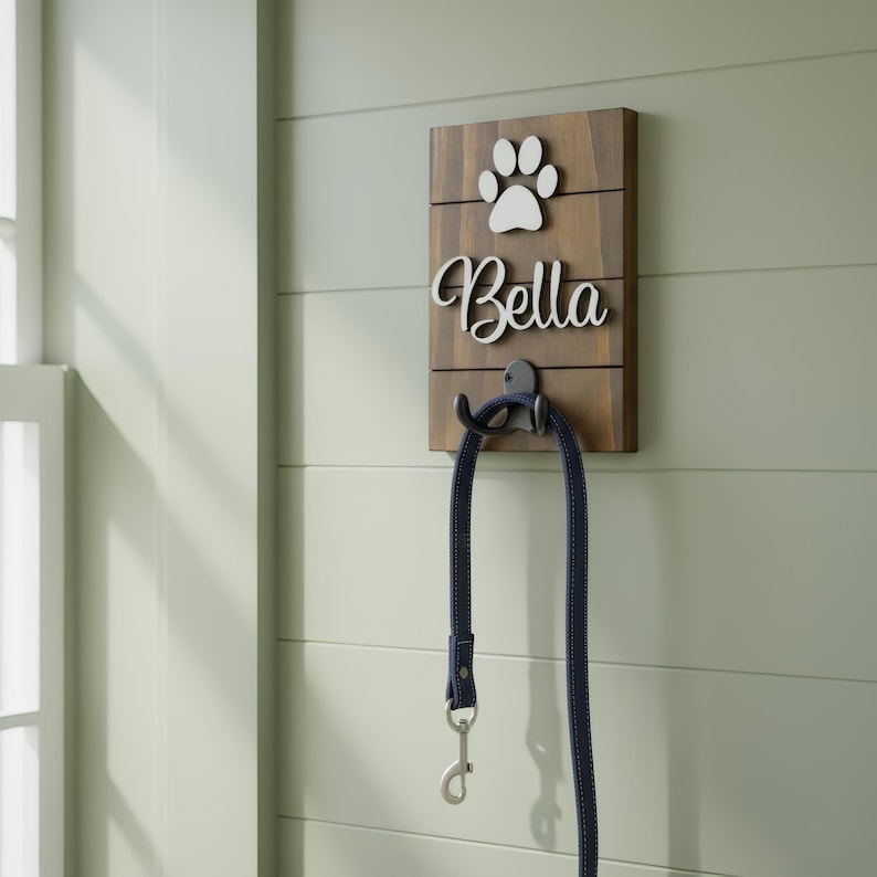 May include: A rectangular wooden wall hook featuring a dark brown finish with horizontal slats. The board displays a white paw print icon above the white script text Bella. A black metal hook is attached, holding a dark blue pet leash.