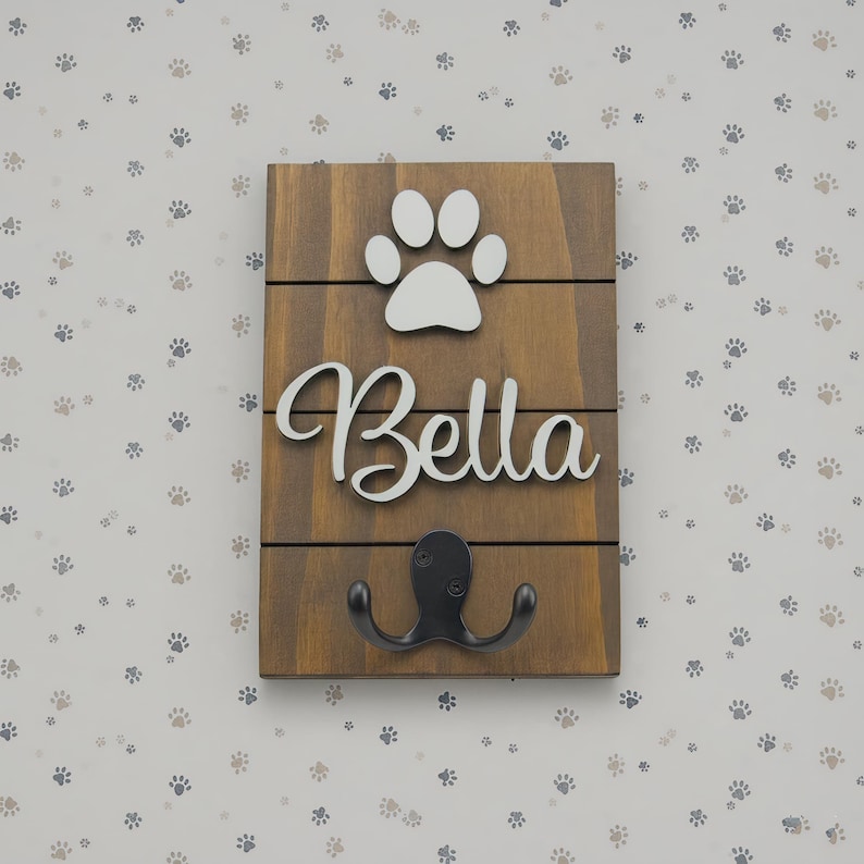 May include: A rectangular wooden wall hook featuring a dark brown stained finish with horizontal slats. The piece displays a white cutout of a paw print above the white script text Bella, with a black metal double hook mounted at the bottom.