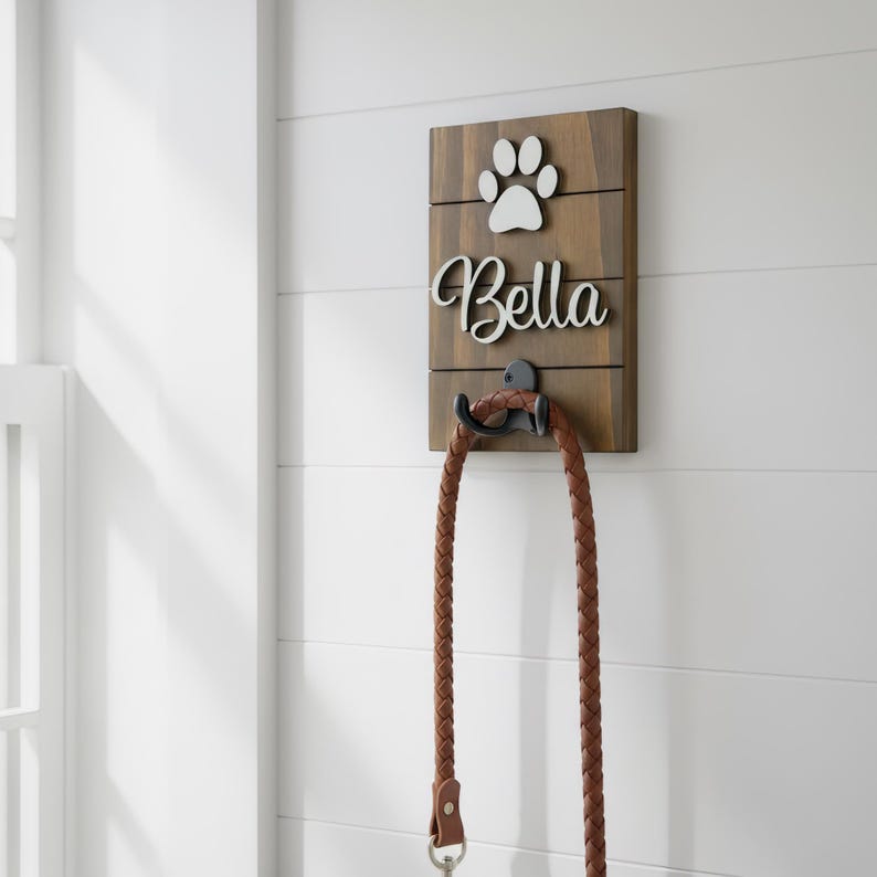 May include: A rectangular wooden wall hook featuring a dark brown finish with horizontal slats. The sign displays a white paw print icon above the white script text Bella. A black metal hook holds a braided brown leather leash hanging downward.