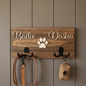 May include: A rectangular wooden wall-mounted rack with a dark brown finish, featuring the names Bodie and Dasha in light-coloured script lettering, a central paw print cutout, and two black metal double hooks holding a leash and a waste bag.
