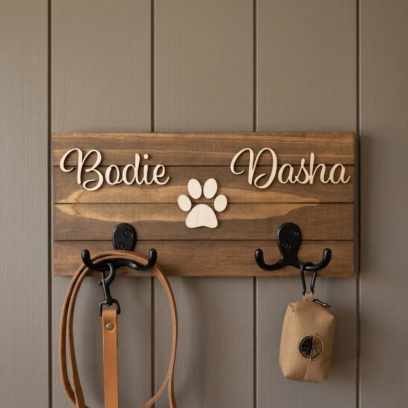 May include: A rectangular wooden wall-mounted rack with a dark brown finish, featuring the names Bodie and Dasha in light-coloured script lettering, a central paw print cutout, and two black metal double hooks holding a leash and a waste bag.