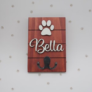 May include: A rectangular wooden wall hook plaque featuring a reddish-brown stained finish with horizontal slats. The plaque displays a white cutout of a paw print above the white script text Bella and a black metal double hook at the bottom.