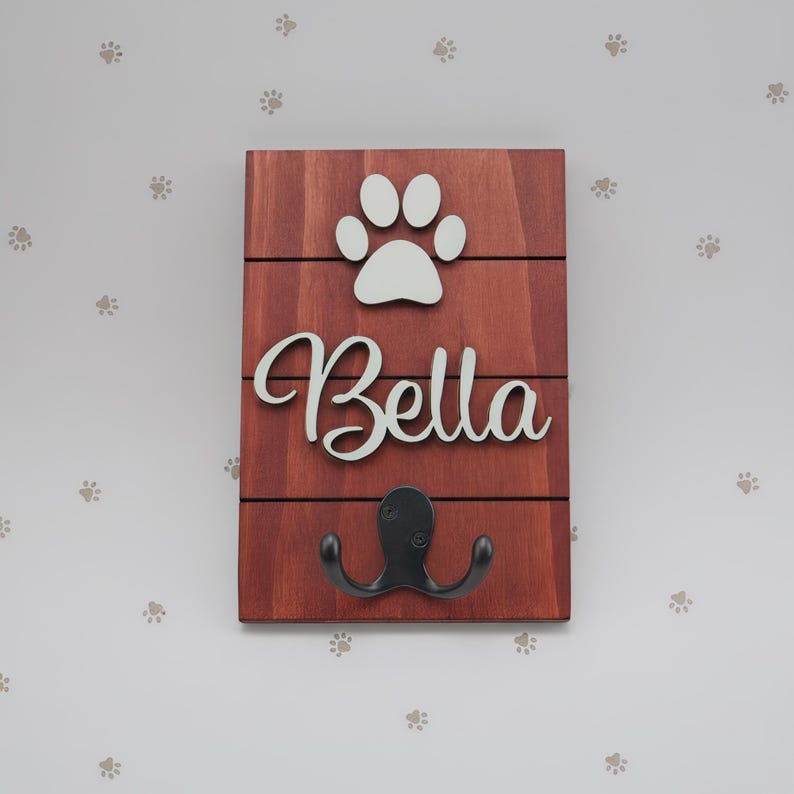 May include: A rectangular wooden wall hook plaque featuring a reddish-brown stained finish with horizontal slats. The plaque displays a white cutout of a paw print above the white script text Bella and a black metal double hook at the bottom.