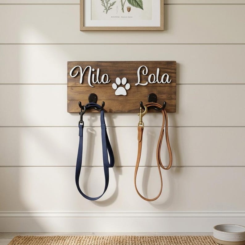 May include: A rectangular dark brown wooden wall-mounted leash holder featuring the white cutout names Nilo and Lola separated by a white paw print. Two black metal hooks are attached to the wood, holding one navy blue lead and one tan lead.