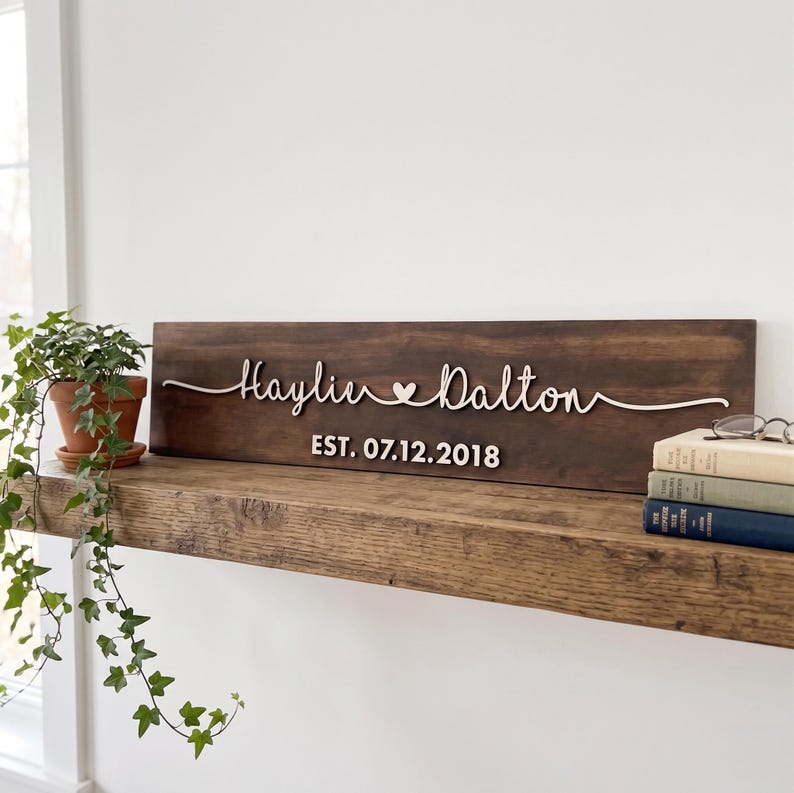 May include: A rectangular dark brown wooden sign featuring white 3D lettering that reads Haylie and Dalton with a small heart symbol in between. Below the names, the text reads EST. 07.12.2018 in white. The sign is displayed on a wooden shelf.