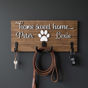 May include: A rectangular wooden wall-mounted key and leash holder with a dark brown finish. It features white 3D lettering that reads "home sweet home" above the names "Peter" and "Lexie" with a white paw print icon. Three black metal hooks.