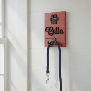 May include: A rectangular red wooden wall-mounted leash holder featuring a black paw print icon and the name Bella in black script lettering. A black metal hook is attached to the centre, holding a dark blue dog leash with a silver metal clasp.