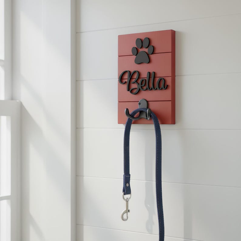 May include: A rectangular red wooden wall-mounted leash holder featuring a black paw print icon and the name Bella in black script lettering. A black metal hook is attached to the centre, holding a dark blue dog leash with a silver metal clasp.
