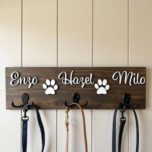 May include: A dark brown wooden wall-mounted rack featuring three black metal hooks. The board displays the white script names Enzo, Hazel, and Milo, separated by two white paw print cutouts. Three pet leashes hang from the metal hooks.