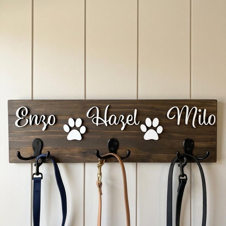 May include: A dark brown wooden wall-mounted rack featuring three black metal hooks. The board displays the white script names Enzo, Hazel, and Milo, separated by two white paw print cutouts. Three pet leashes hang from the metal hooks.