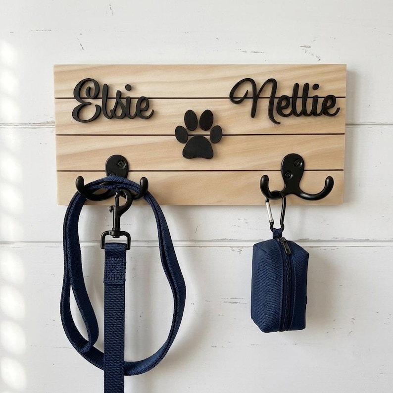 May include: A rectangular light-coloured wooden wall-mounted rack featuring two black metal hooks. The names Elsie and Nettie are written in black script lettering above a black paw print icon. A navy blue leash and a navy blue waste bag holder hang.