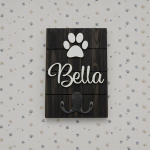 May include: A rectangular dark wood wall plaque featuring three horizontal planks. A white cutout of a paw print sits above the white script text Bella. A black metal double hook is attached to the bottom section of the dark wood surface.