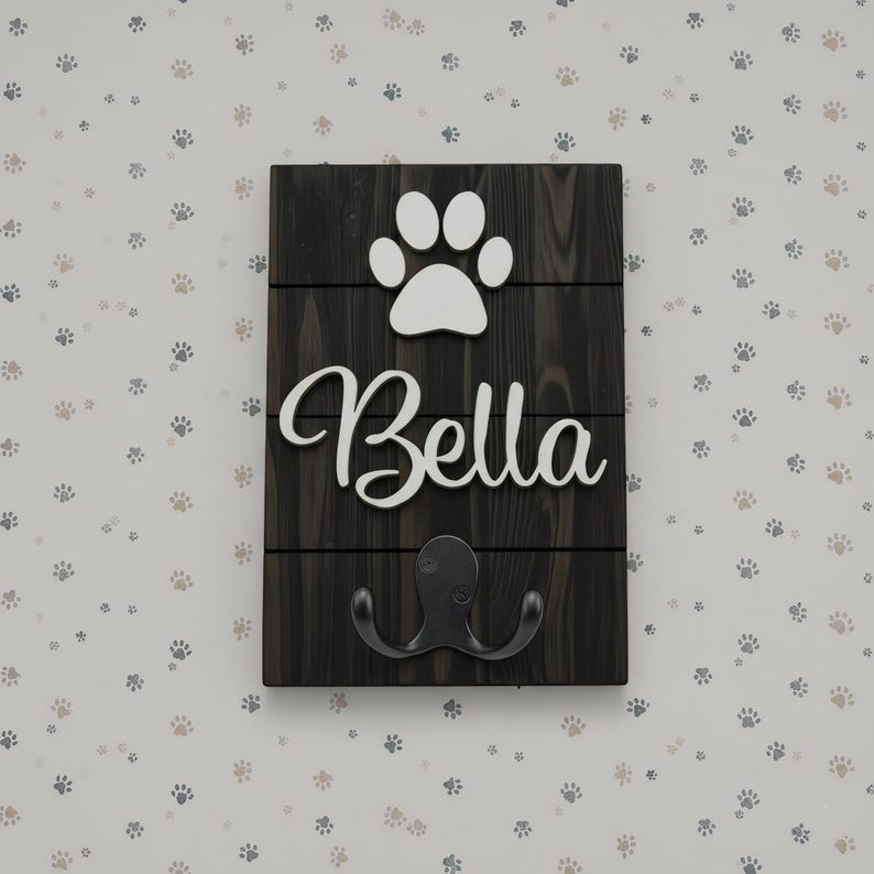 May include: A rectangular dark wood wall plaque featuring three horizontal planks. A white cutout of a paw print sits above the white script text Bella. A black metal double hook is attached to the bottom section of the dark wood surface.
