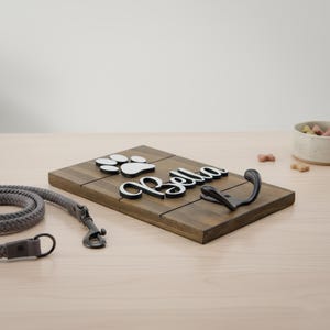 May include: A rectangular dark brown wooden wall hook plaque featuring a white paw print cutout and the name Bella in white script lettering. A black metal hook is attached to the right side of the wood panel, which has horizontal grooves.
