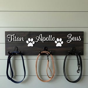 May include: A dark brown wooden wall-mounted rack featuring three black metal hooks holding leashes. The board displays the white script names Titan, Apollo, and Zeus, interspersed with two white paw print icons, creating a personalised pet accessory organiser.