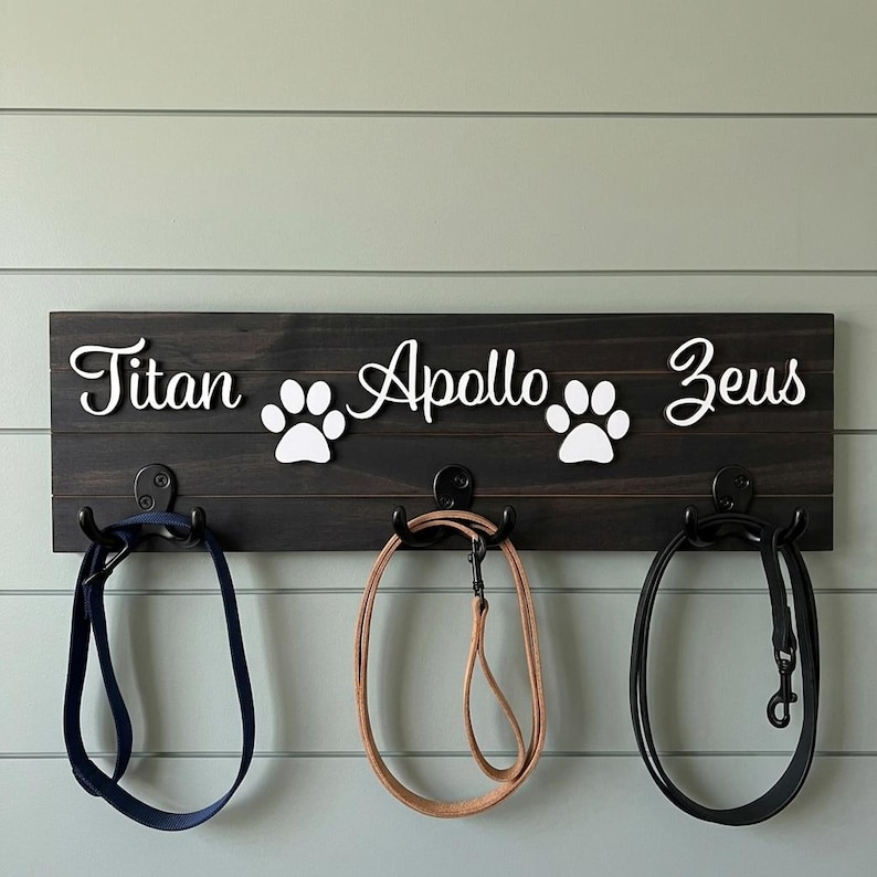 May include: A dark brown wooden wall-mounted rack featuring three black metal hooks holding leashes. The board displays the white script names Titan, Apollo, and Zeus, interspersed with two white paw print icons, creating a personalised pet accessory organiser.