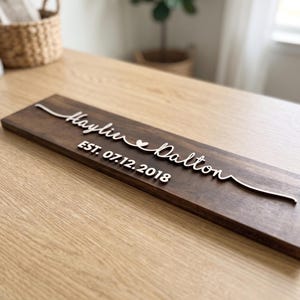 以下が含まれることがあります： Haylie Daltonと書かれた白い浮き彫りの文字が特徴の長方形のダークブラウンの木製看板。名前の間には小さなハートのアイコンがあり、そのすぐ下にEST. 07.12.2018というテキストがはっきりとしたフォントで配置されています。