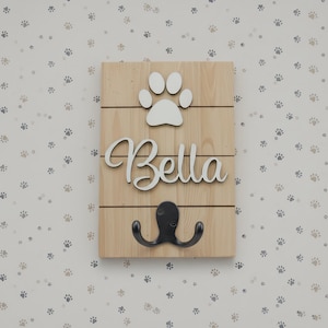 May include: A rectangular light-coloured wooden wall plaque featuring a white paw print cutout at the top, the name Bella in white cursive script lettering in the centre, and a black metal double hook mounted at the bottom for hanging items.