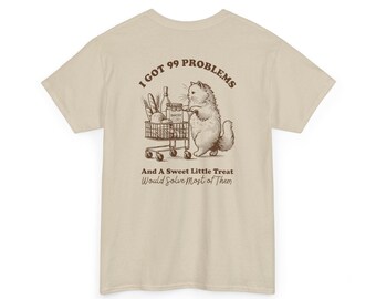 以下が含まれることがあります： ベージュのTシャツに、食料品でいっぱいのショッピングカートを押す猫の茶色のグラフィックが描かれています。Tシャツには「I got 99 problems and a sweet little treat would solve most of them」というテキストが印刷されています。