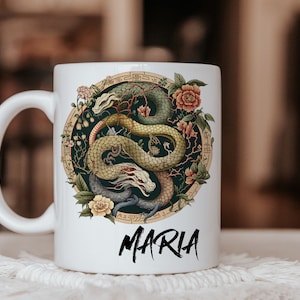 Könnte beinhalten: Weiße Keramiktasse mit einer detaillierten Illustration von zwei verschlungenen Drachen in Grün-, Gold- und Grautönen, umgeben von floralen Elementen. Der Name "MARIA" steht in schwarzer Schrift unten.