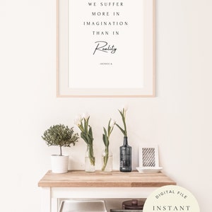 Pu&ograve; includere: Una stampa incorniciata con sfondo bianco e testo nero che recita "We suffer more in imagination than in Reality - Seneca". La stampa &egrave; appesa a una parete bianca sopra un tavolo di legno con fiori bianchi in vasi di vetro, una piccola pianta in vaso e un vassoio decorativo.