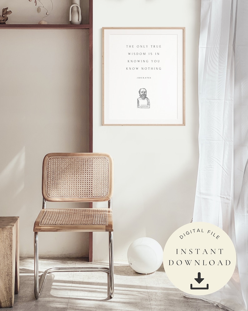 May include: A printable digital download of a quote by Socrates, "The only true wisdom is in knowing you know nothing." The quote is printed in black text on a white background with a black and white illustration of Socrates. The quote is framed in a light wood frame and is hanging on a white wall. A wicker chair and a white ball are in the foreground.