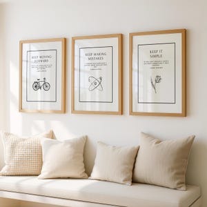 K&ouml;nnte beinhalten: Drei gerahmte Kunstdrucke mit inspirierenden Zitaten. Die Rahmen sind aus hellem Holz, mit wei&szlig;em Hintergrund und schwarzen Texten und Illustrationen. Darunter liegen drei Kissen auf einer wei&szlig;en Bank.