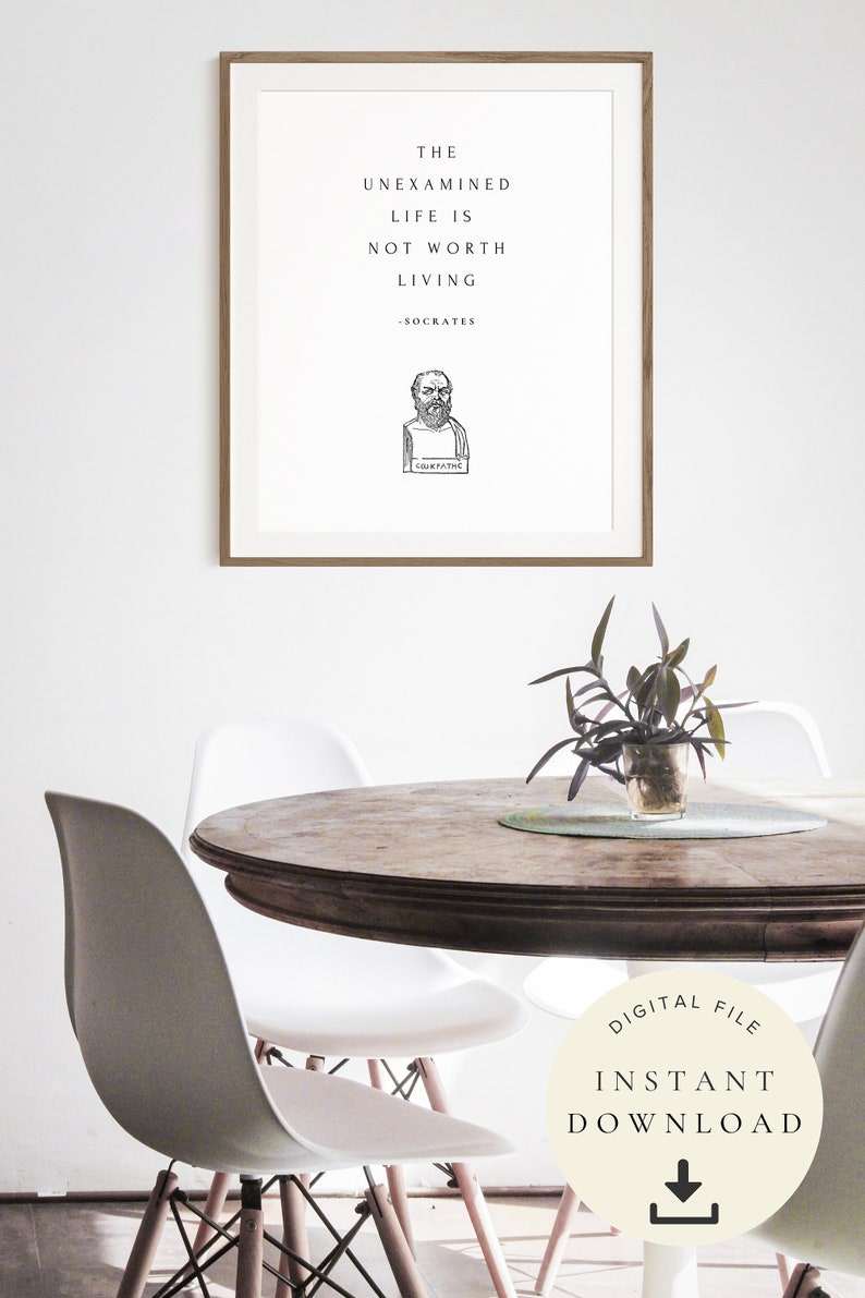 May include: A framed print with a white background and black text that reads "The unexamined life is not worth living - Socrates". A black and white illustration of Socrates is below the text. The print is hanging on a white wall above a wooden table with two white chairs. A small plant is on the table.
