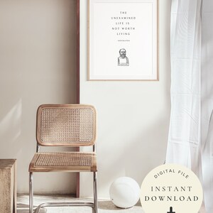 May include: A framed print with a quote by Socrates: "The unexamined life is not worth living." The print is hanging on a white wall with a wicker chair and a white ball in the foreground.