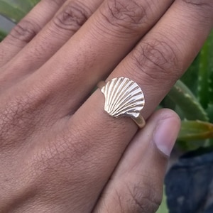 Könnte beinhalten: Ein goldfarbener Ring mit einem Muscheldesign. Die Muschel des Rings ist mit radialen Linien detailliert, und das Band ist glatt. Der Ring wird auf einem Finger vor einem verschwommenen grünen Hintergrund gezeigt.