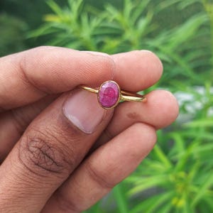 Op de afbeelding: Een gouden ring met een levendige, ovale, rode edelsteen. De ring heeft een slanke, gepolijste gouden band en de edelsteen is gezet in een eenvoudige bezel. De achtergrond is een wazig beeld van groen gebladerte.