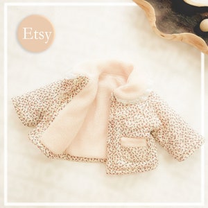 Op de afbeelding: Een wit en roze babyjasje met een bloemenprint en een witte kantversiering. Het jasje is gevoerd met zacht, roze fleece.