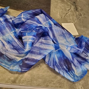 Può includere: Sciarpa leggera con motivi blu e bianchi. Il tessuto presenta un motivo ad acquerello con accenti blu scuro, creando un design dinamico e artistico. La sciarpa è drappeggiata, mostrando la sua consistenza e la sua caduta.