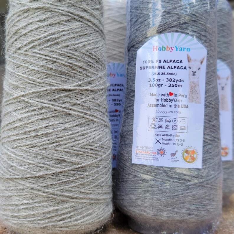Pode incluir: Um cone de fio de alpaca superfina cinza-claro da HobbyYarn. O r&oacute;tulo apresenta uma ilustra&ccedil;&atilde;o de alpaca, o texto 100% FS Alpaca, 100g, 350m e instru&ccedil;&otilde;es de cuidado. O fio est&aacute; envolto em pl&aacute;stico transparente, com textura macia.