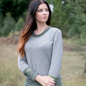 May include: A gray long-sleeved sweater with green and blue trim around the neckline and cuffs. The sweater has a ribbed texture and a crew neck. The model is wearing the sweater outdoors in front of a blurred green background.